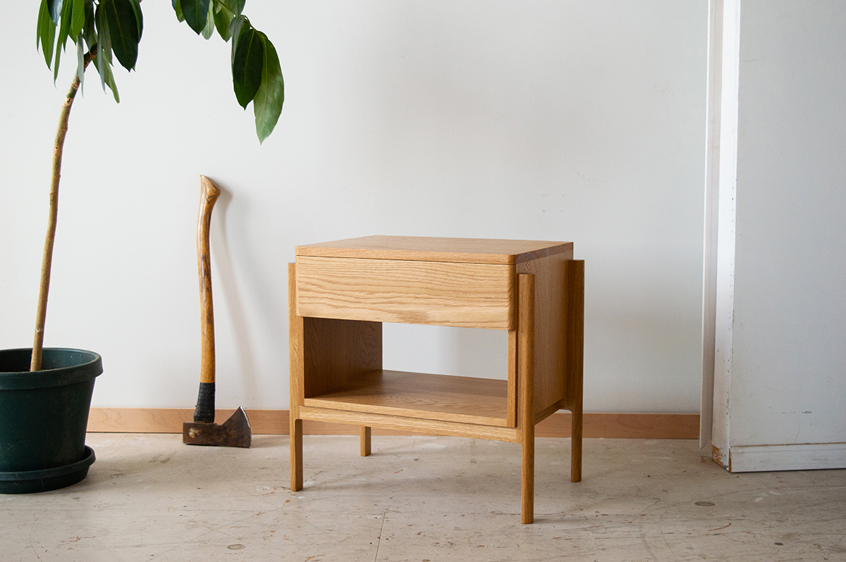 A well-crafted nightstand sits in a space next to a plant.