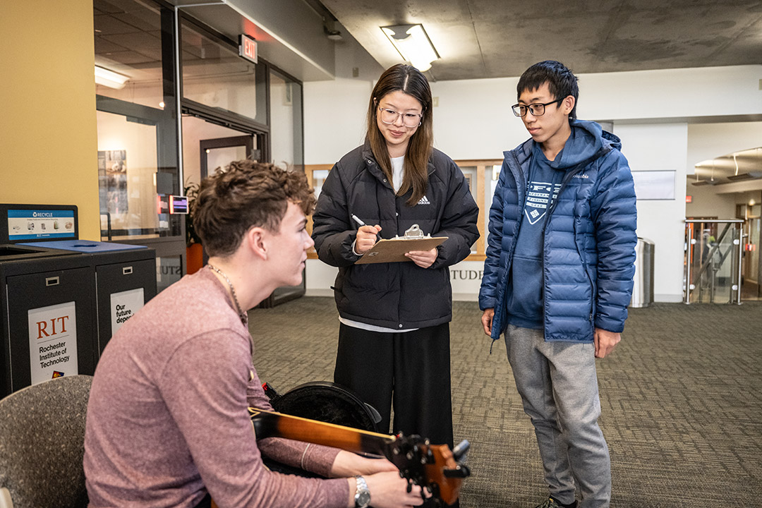 two college students standing, one with a clipboard, next to another student who is seated.