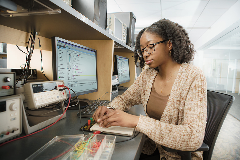 An RIT student, Erica Coles wearing a tan sweater and inserting wires into a computer bread board at a desk with a desktop computer.