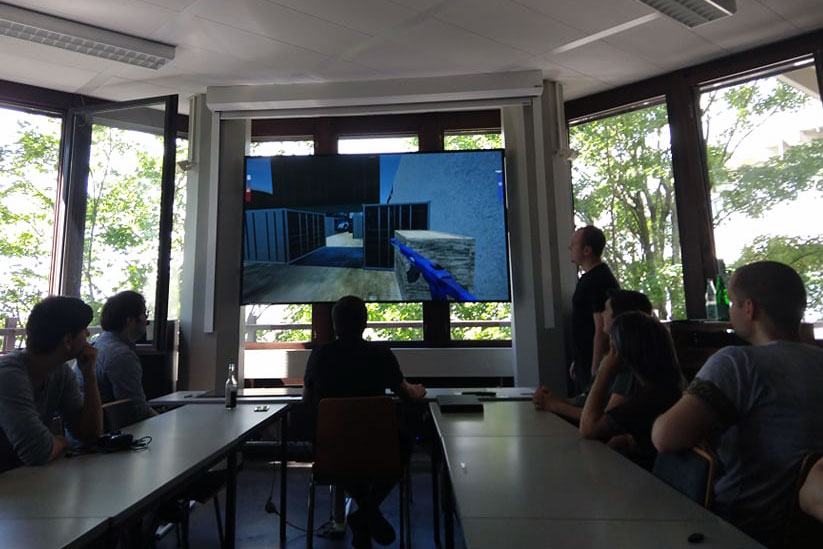 Students sit at two long tables facing large TV screen.