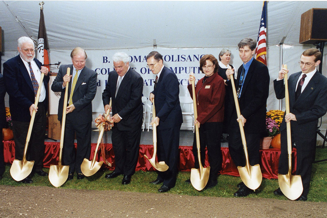 seven people lined up holding golden shovels for a ceremonial groundbreaking.