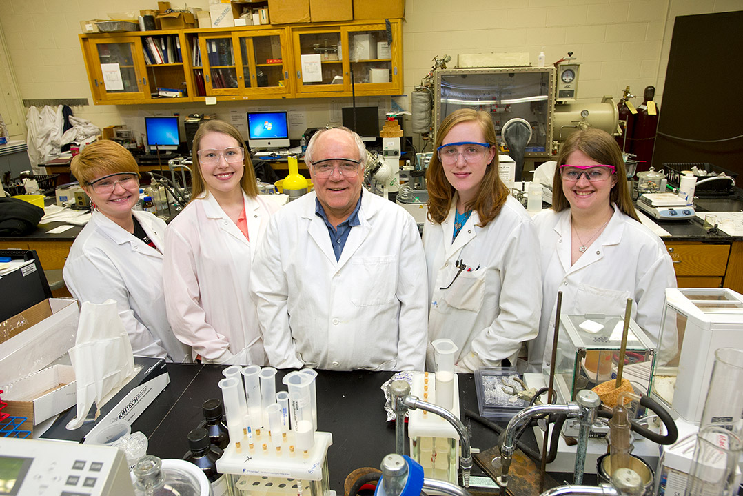 Professor and students in lab coats standing near table of equipment.