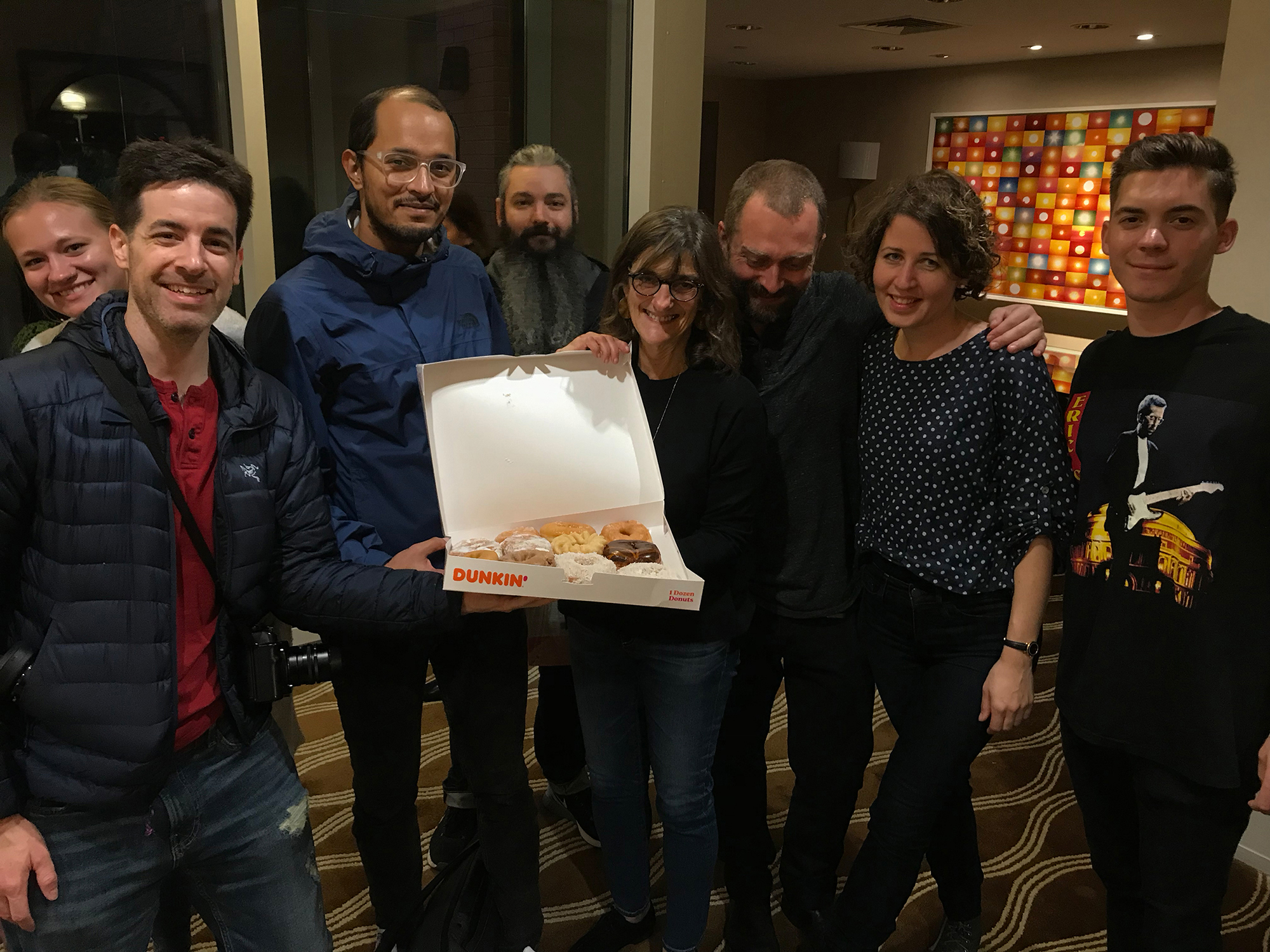A group of people gather around a box of doughnuts.
