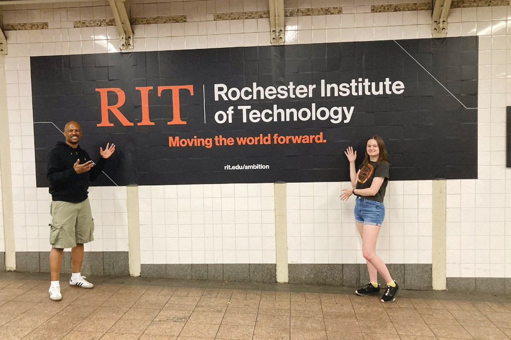 two people standing next to an ad hanging on a subway wall that features one person's picture.