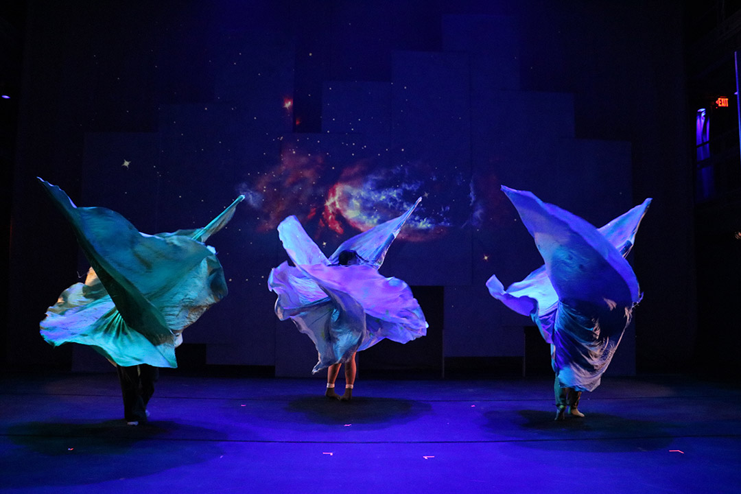 three dancers on a stage rotating green, purple, and blue fabrics in the air.