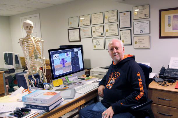 professor sitting at desk facing a model of a human skeleton.