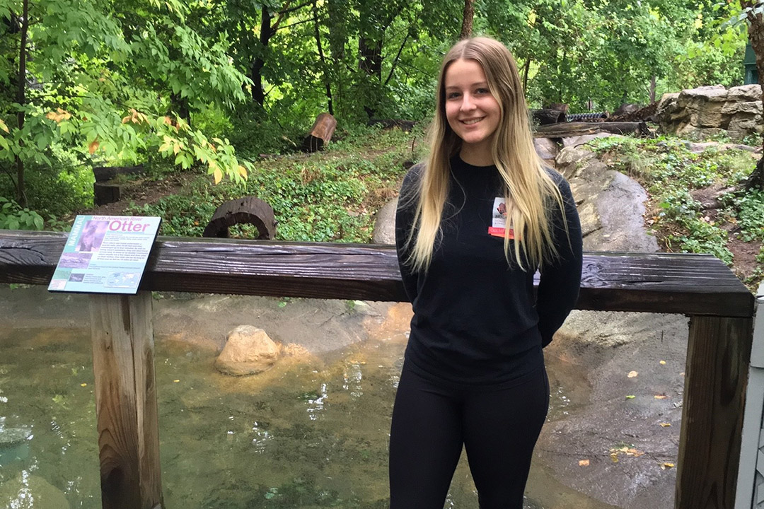 student researcher standing outside the river otter exhibit at a zoo.