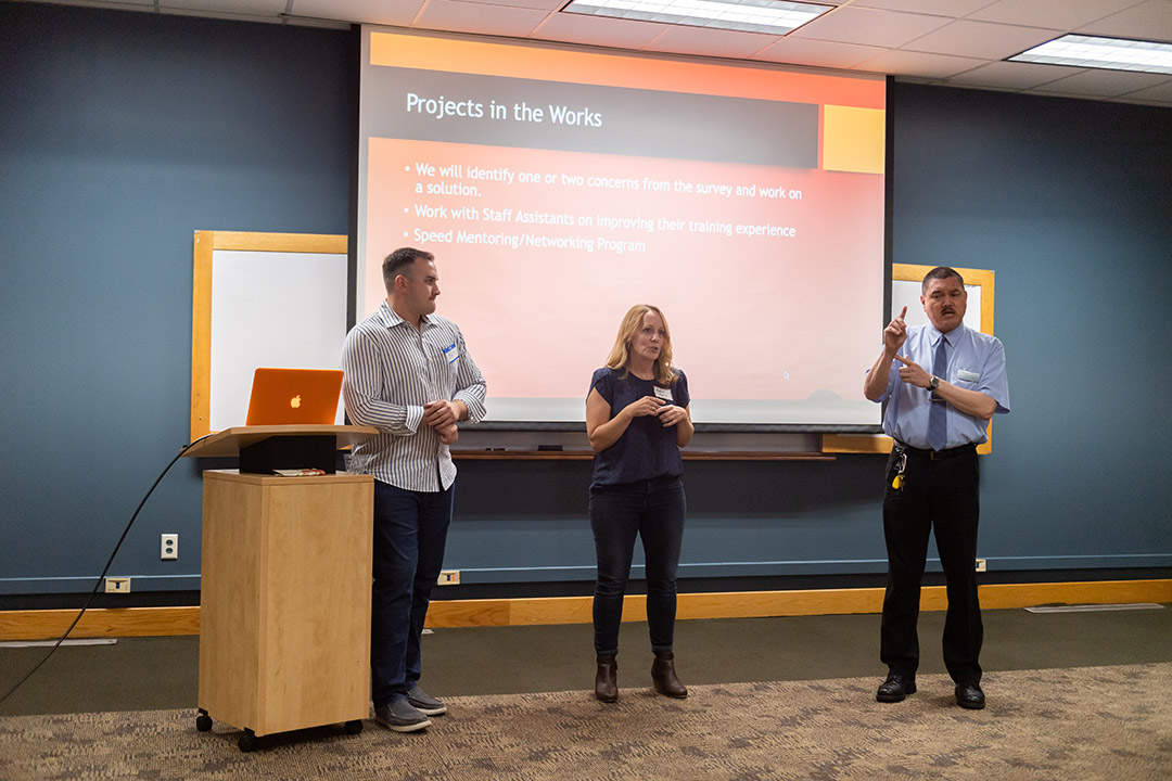 three people, one using sign language, standing in a room giving a presentation.