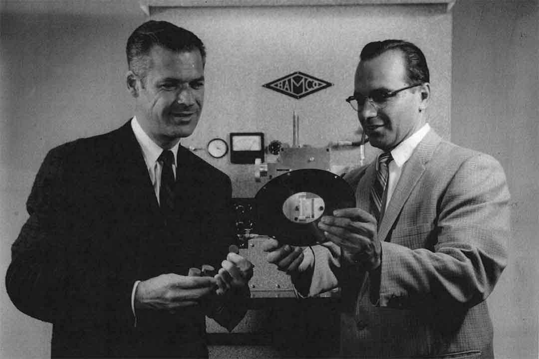 two men looking at a thin slice of a round material in the 1950s.