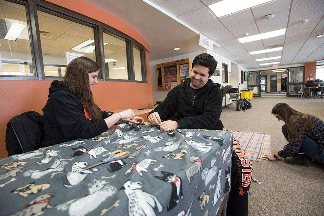 Two students sit at a table and one kneels on the ground. All are making blankets. 