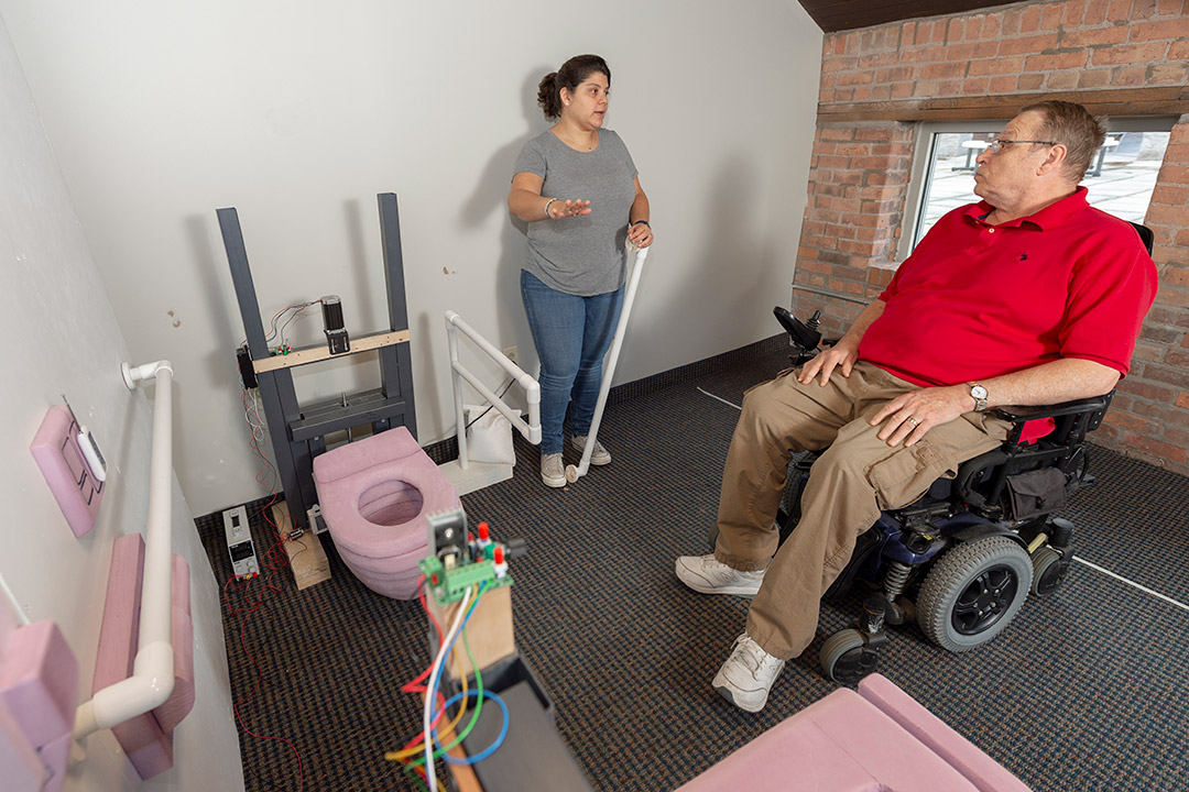 researcher showing prototype of accessbile bathroom to man in wheelchair.