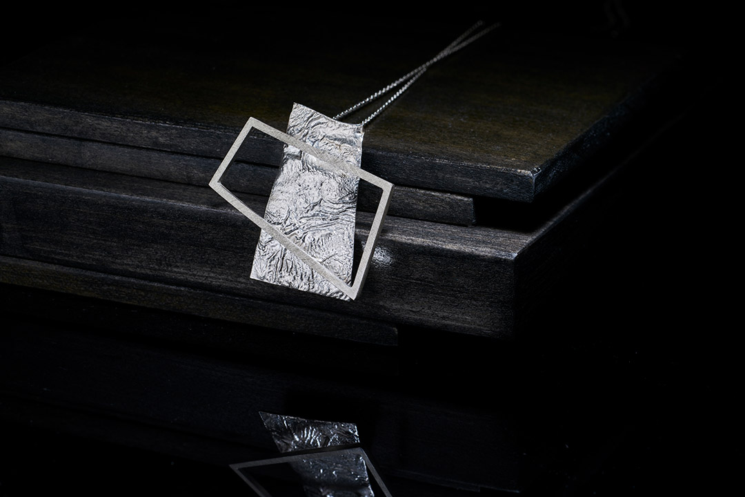 a necklace pendant with an irregular metal rectangle outline over a solid metal rectangle.