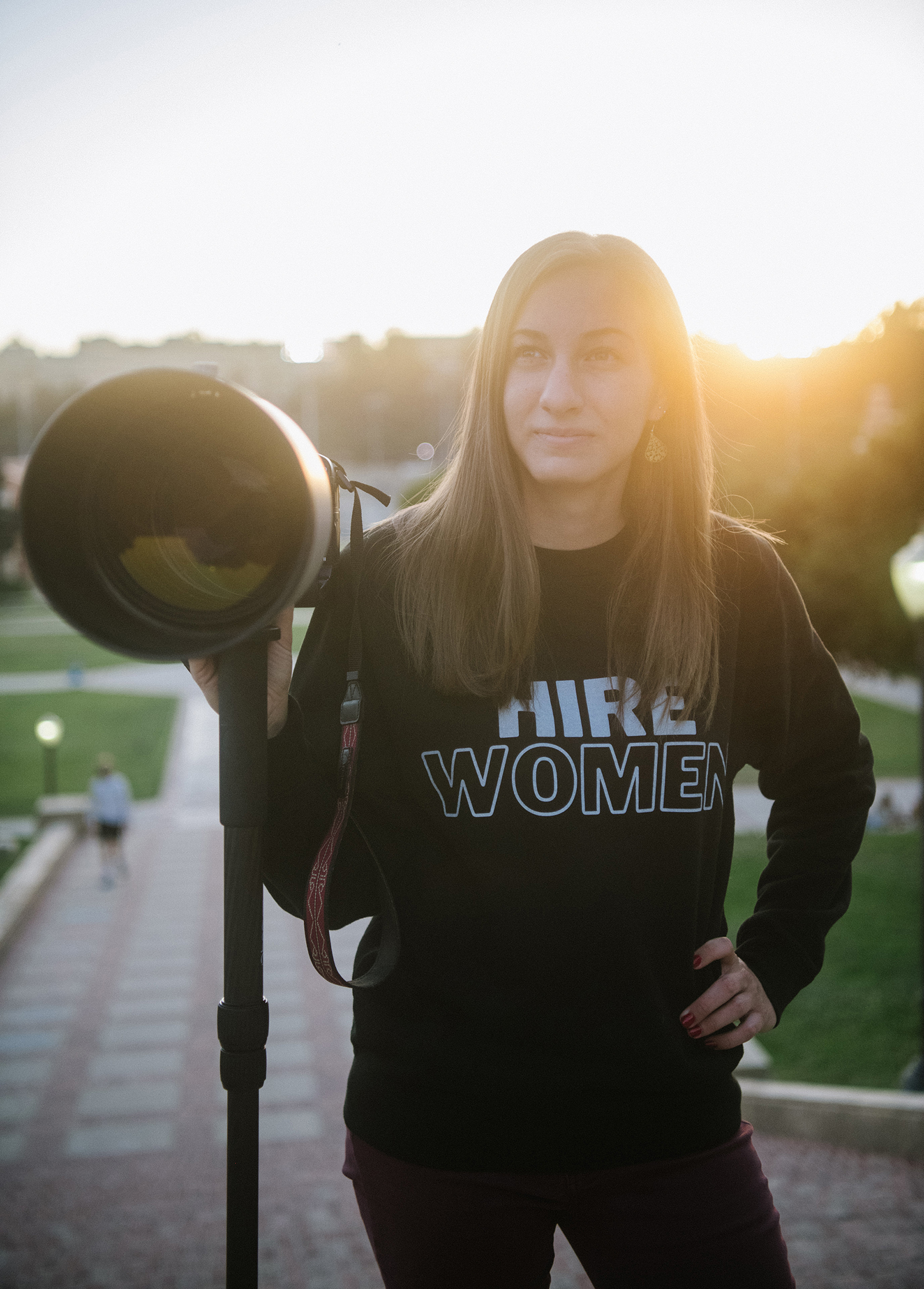 Meg Oliphant poses for a portrait holding a camera and wearing shirt that reads Hire Women.