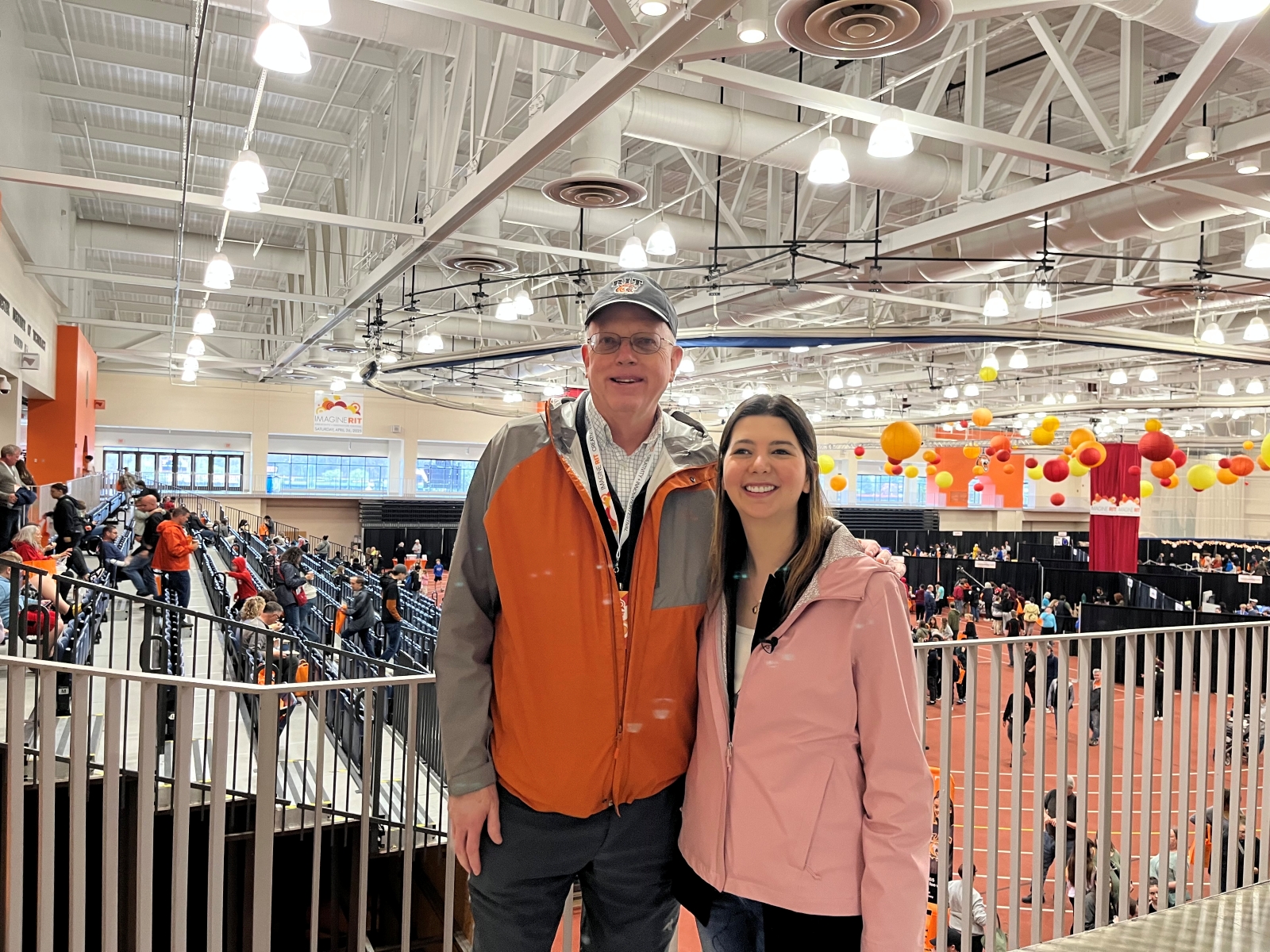 Man in orange jacket poses with woman in pink jacket in a field house with a crowd behind them.