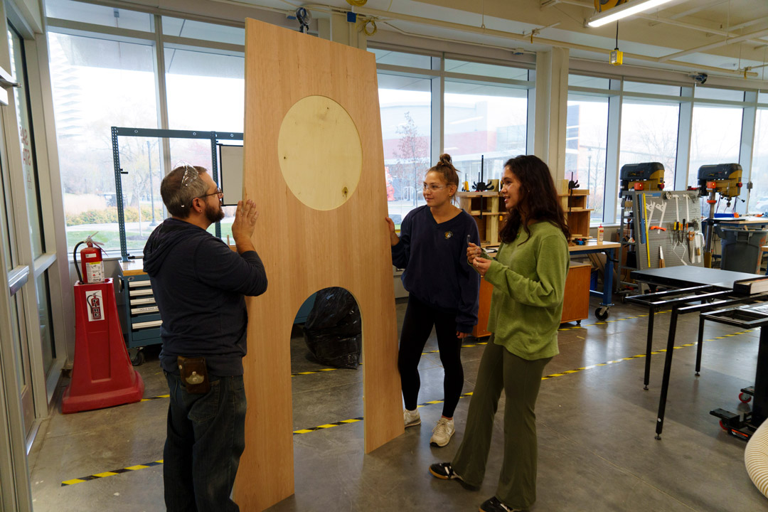Two students and a faculty member stand by and look at a large wood cutout of a trophy.