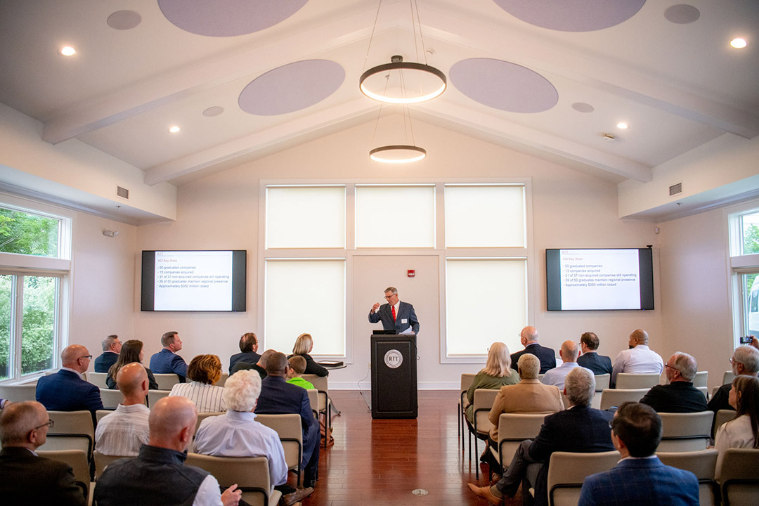 person standing at a podium at the head of a room of about two dozen people.