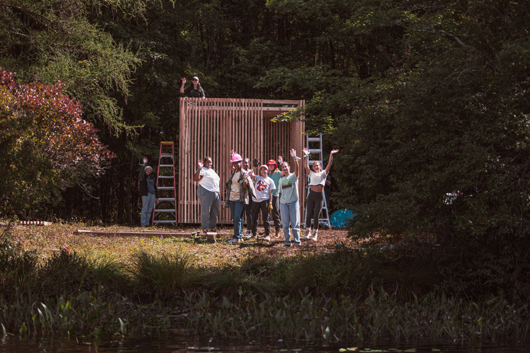 A group stands in a wooded area in front of a small stucture.