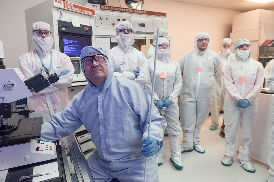 Robert Pearson sits in the MicroE clean room with 6 of his students. All of them are in clean room jump suits. Pearson operates a machine on the left side of the image while the students look on. Pearson and the students are focused on something behind the photographer, so they are all looking toward the viewer.
