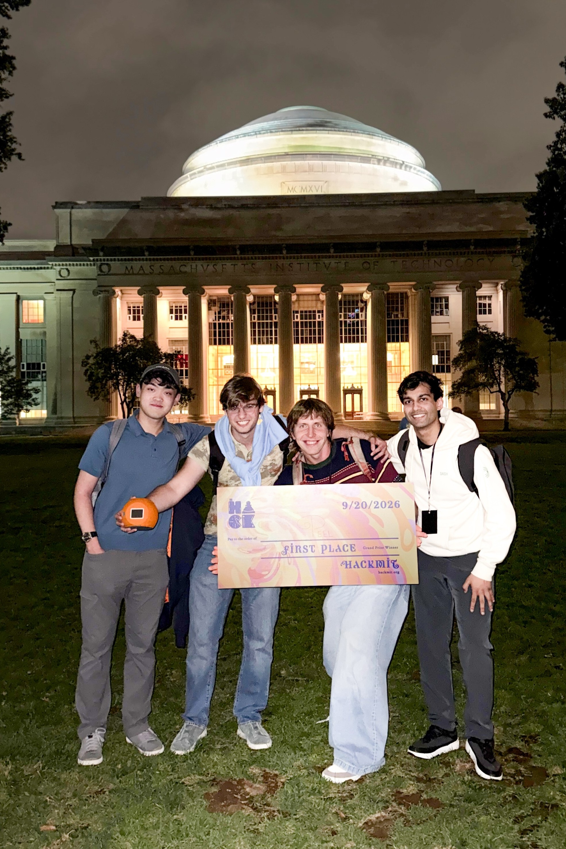 Four young men stand in front of a white domed building with a giant check that states they won first place at Hack M.I.T.