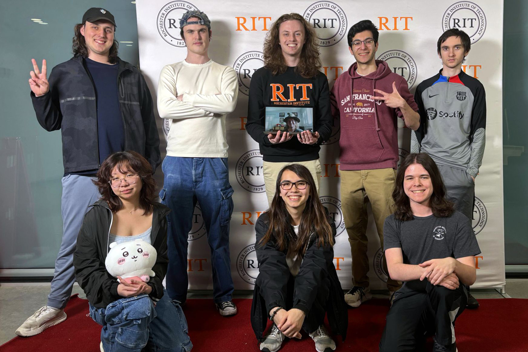 A group of students stands and kneels in front of a backdrop with the RIT logo