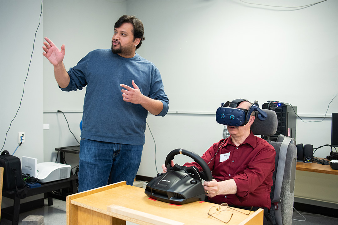 Man wears VR goggles while sitting at table holding a steering wheel.