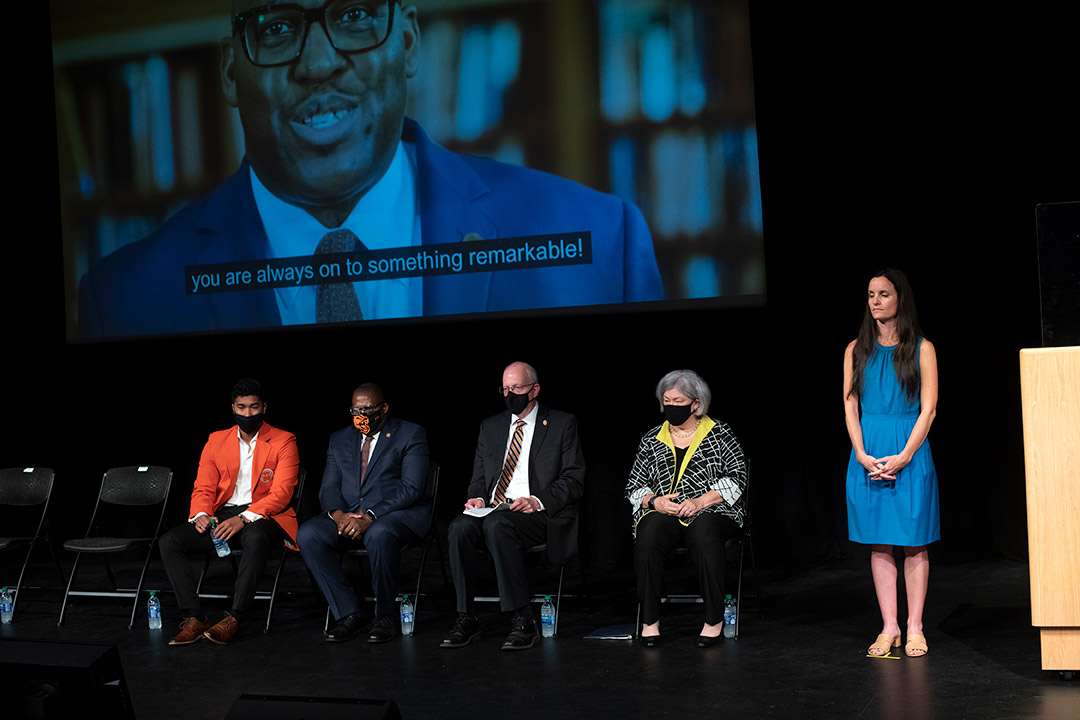 four people sitting and one person standing on a stage while a video plays on a screen in the background.