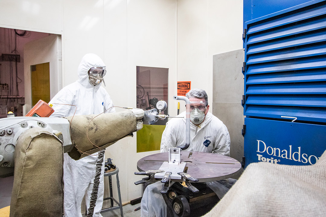 two researchers wearing clean suits looking at a robotic arm.