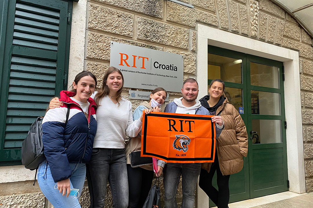 five students in Dubrovnik, Croatia, standing outside a campus building holding an RIT banner.