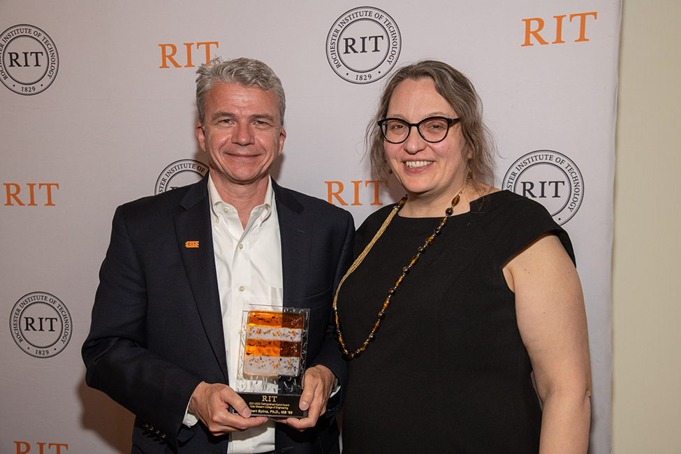 Two people standing and smiling. The person on the left is holding a glass award.