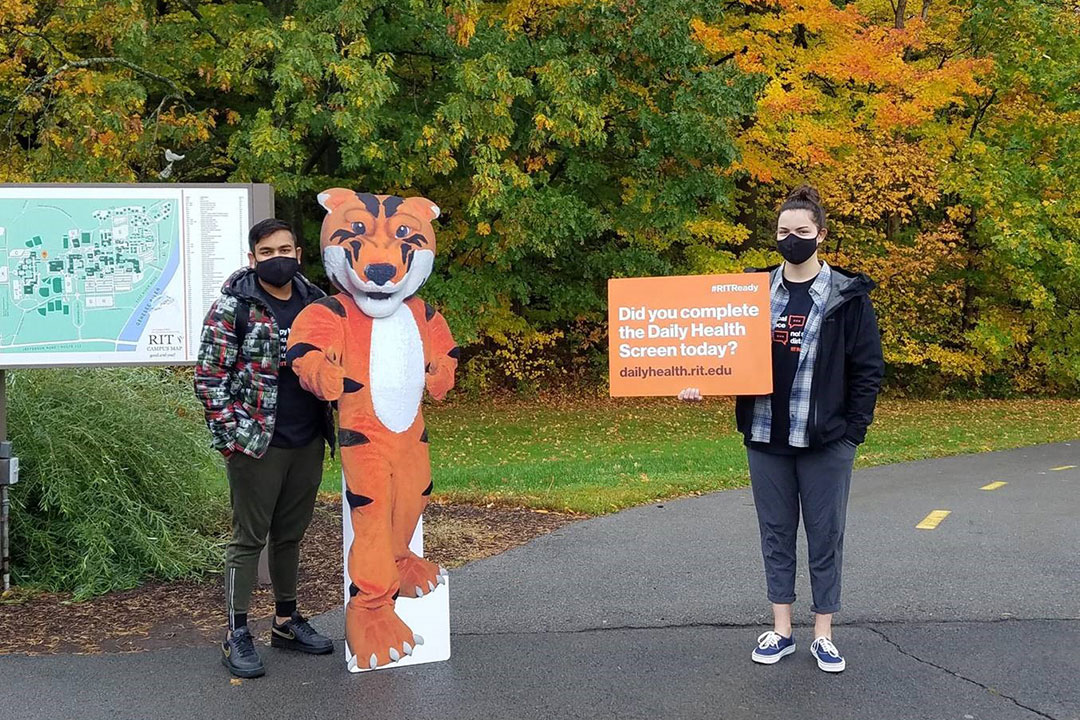 two students with a life-size cardboard cutout of tiger mascot and sign about daily health screen.