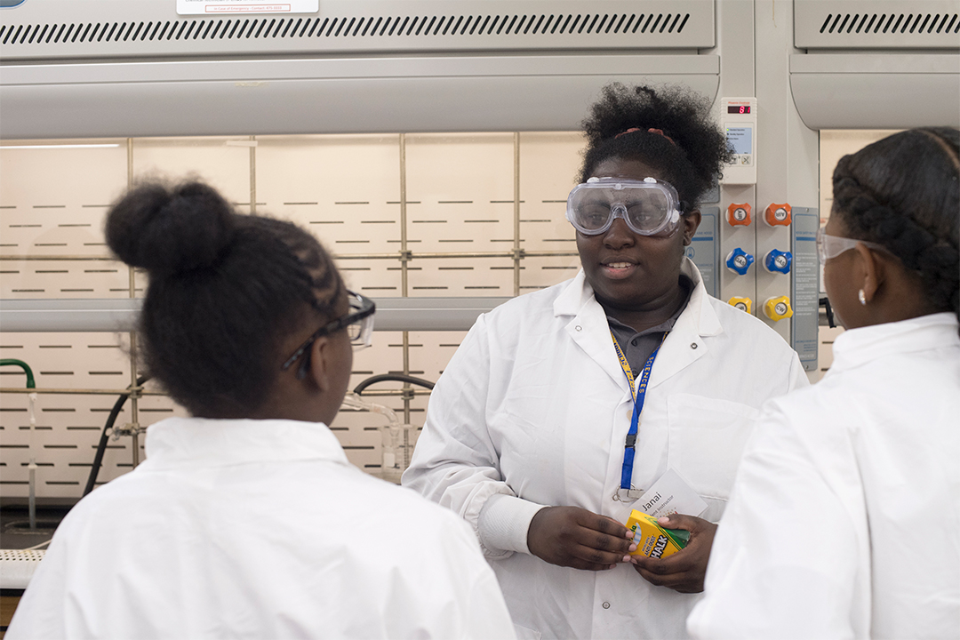 Students in lab coats talk to each other.