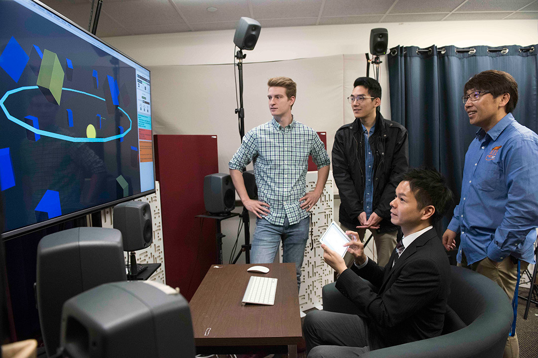 Students and researchers sit and stand around TV screen displaying geometric shapes