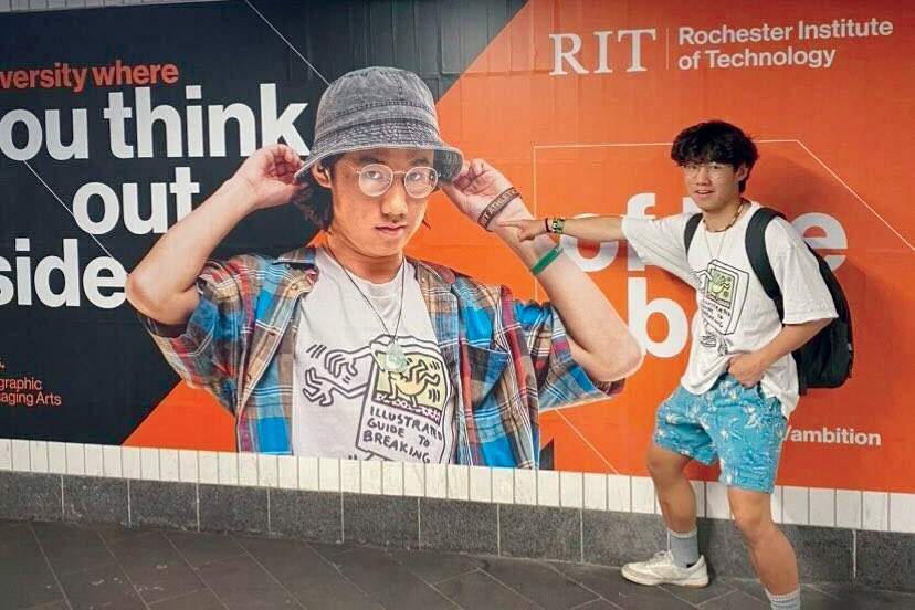 college graduate standing next to and pointing at an ad hanging on a subway wall that features his picture.