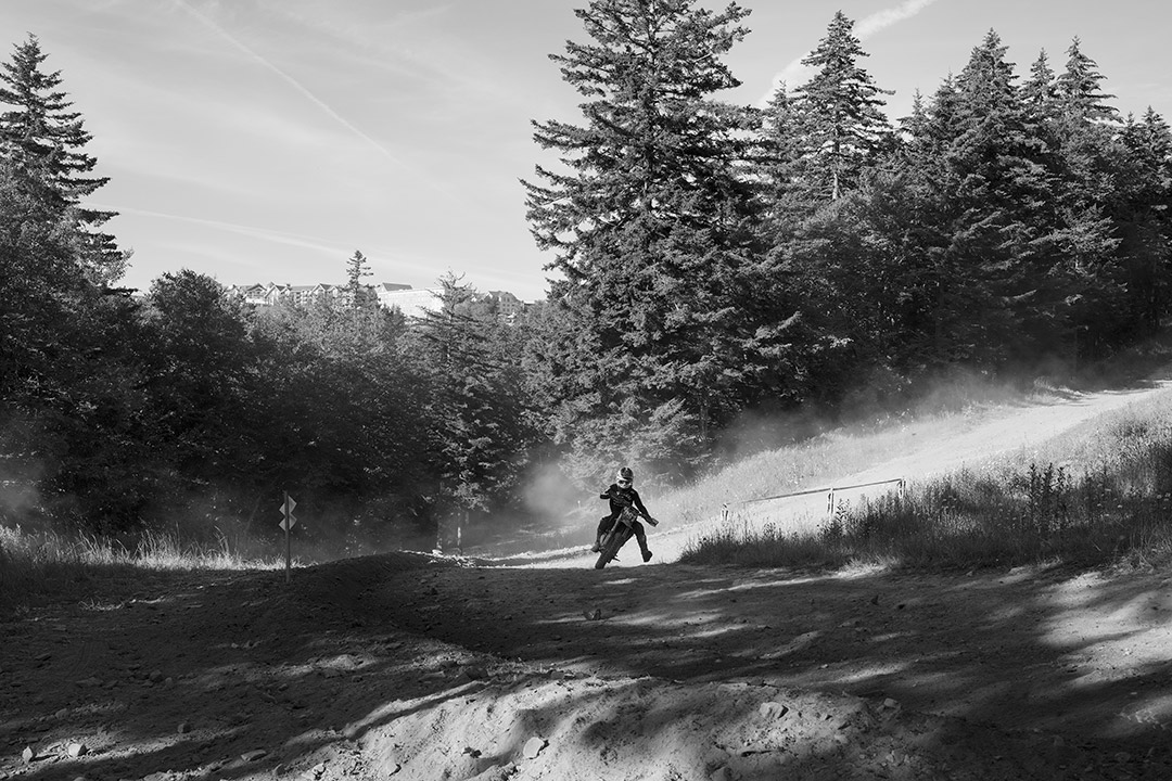 person on a dirt bike on a dirt path with tall evergreen trees in the background.