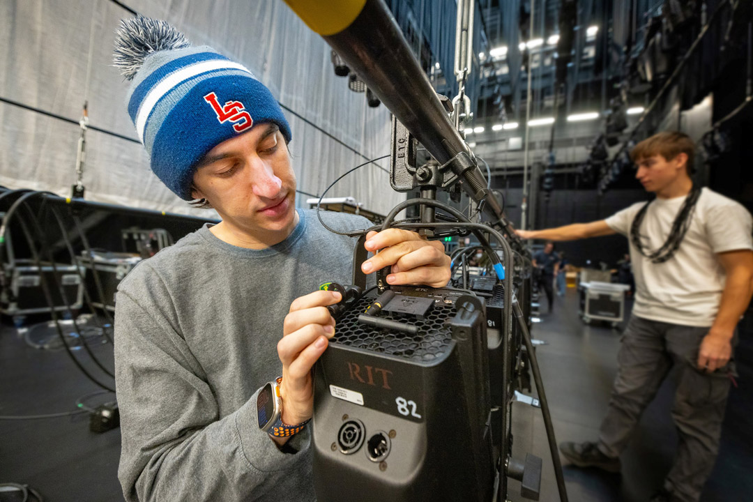 A man installs lights to a large batten on a stage.