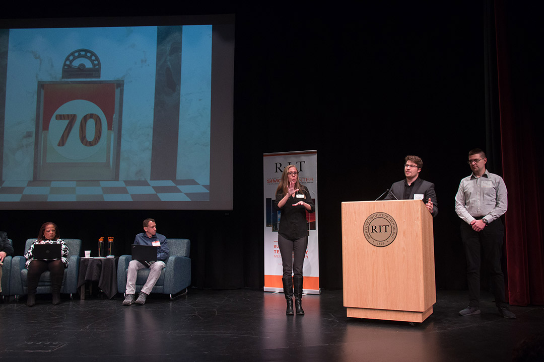 Five people on a stage: 2 seated, 2 speaking at podium and one interpreting in American Sign Language.