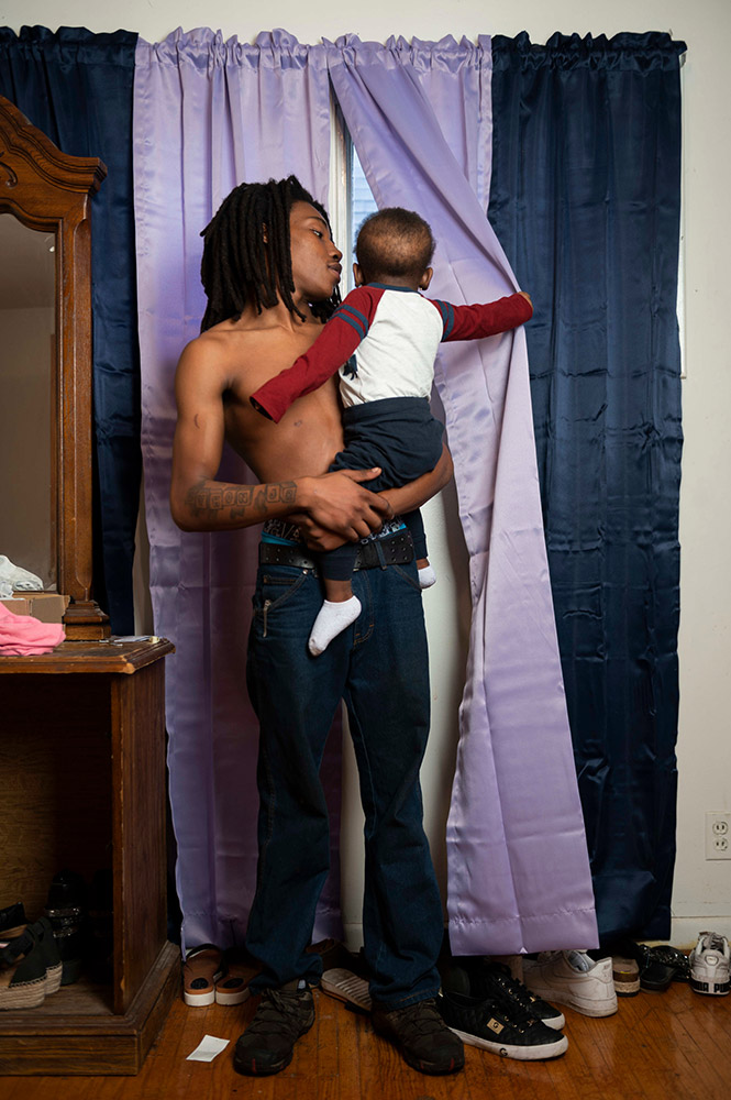 a man holding a toddler, who is pulling back a curtain and looking out a window.