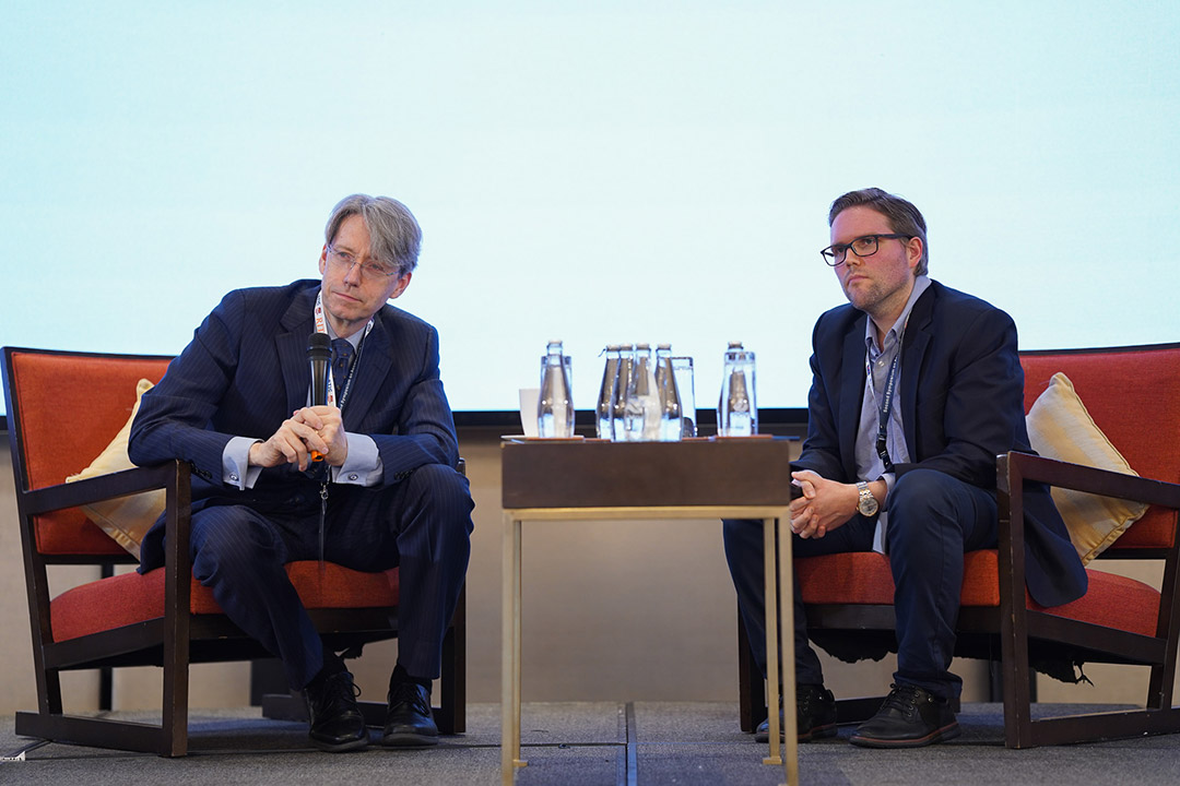 two men sit in chairs on a stage leaning in to listen to someone speak.