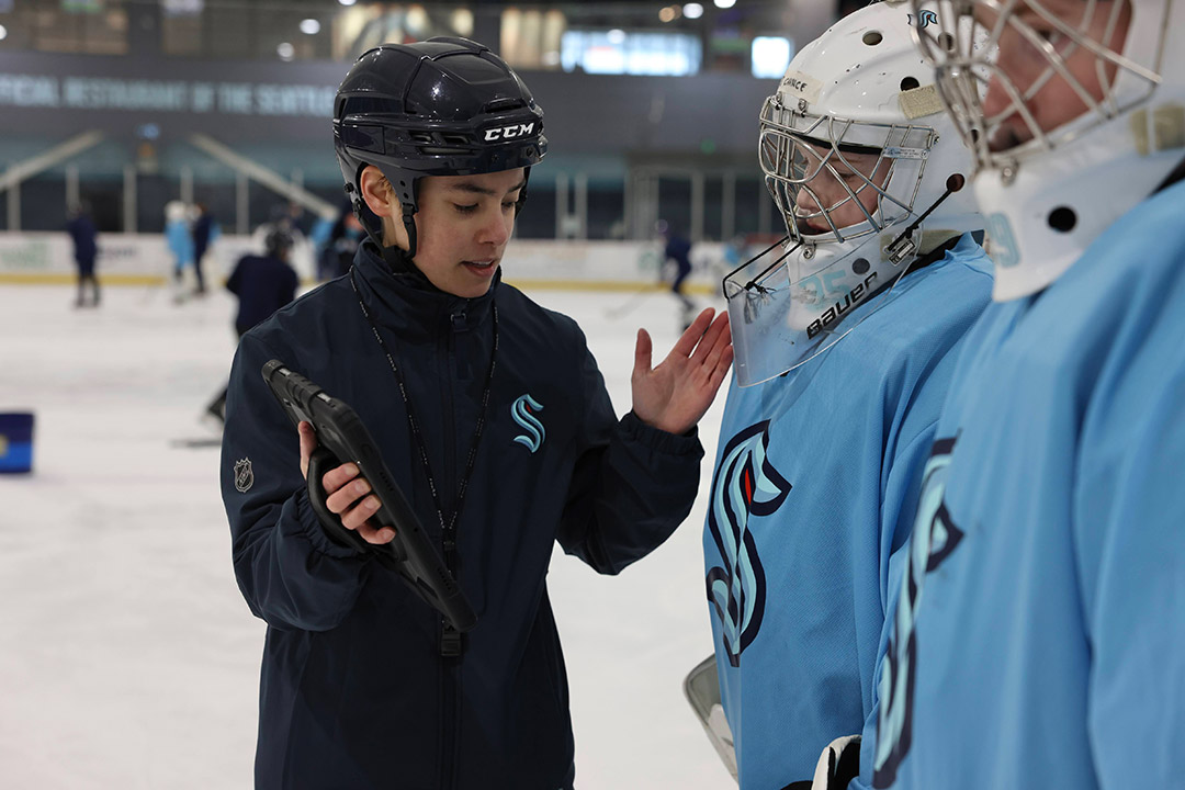 Coach talks to two goalies on the ice while holding a tablet.