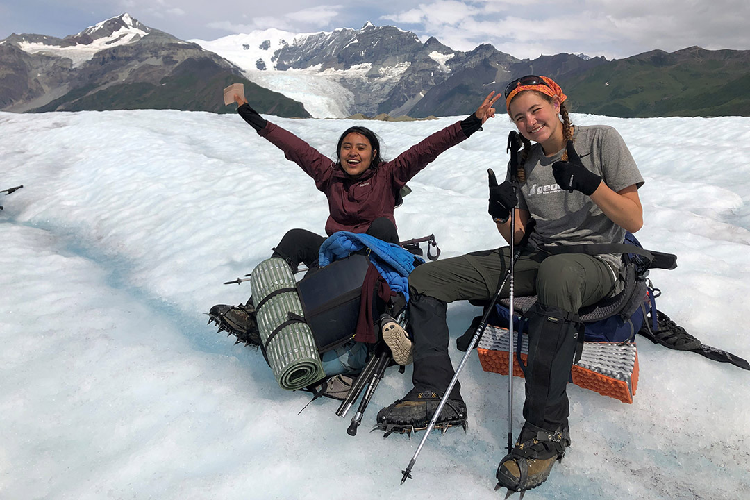 two female students sit on a snowy glacier with winter hiking gear. The female on the left has her hands raised with an excited look on her face, while the female on the left smiles and gives two thumbs up.
