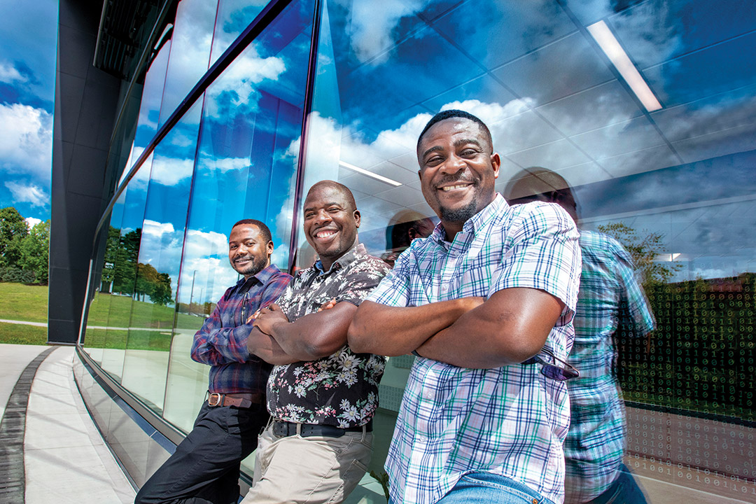 three black students stand in front of a glass front building smiling at the camera.