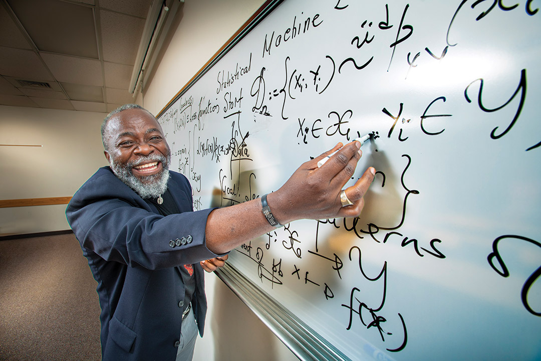 an older black man smiles at the camera as he writes on a whiteboard with marker.