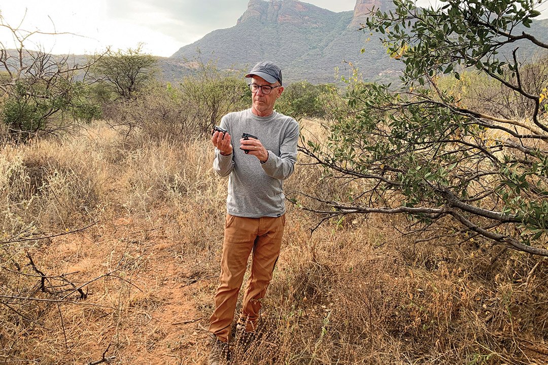 an older man stands holding a small item in his hands in the african plains.