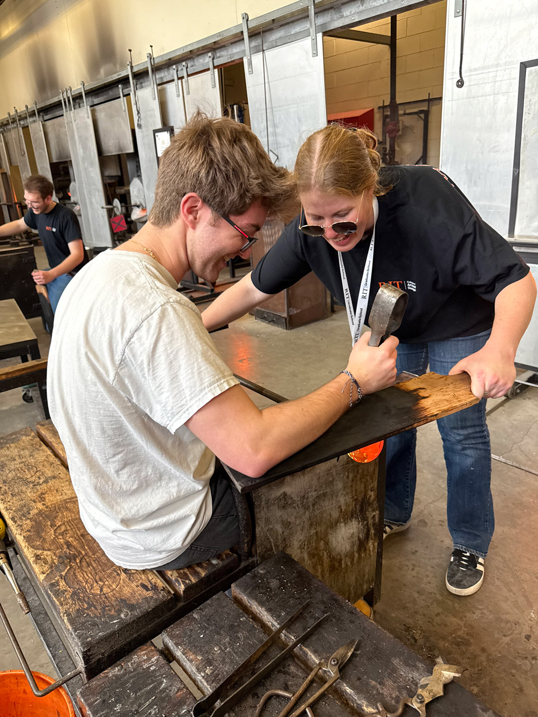 Two students work together manipulating glass in a hot shop workshop