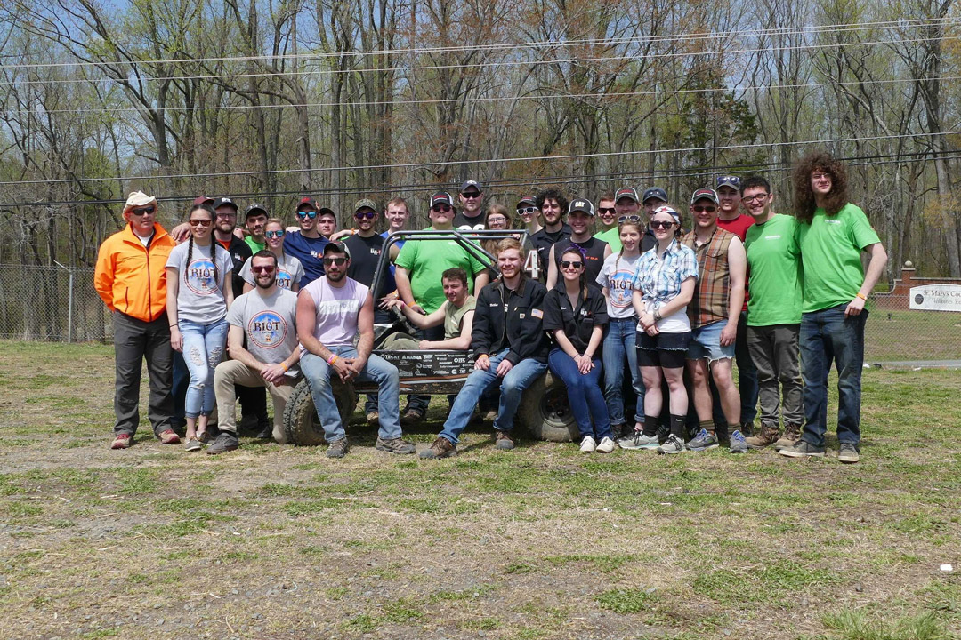 a large group of people gather smiling around an R I T Baja Car.