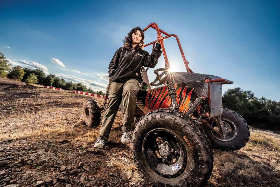 a female student in a black jacket stands next to an R I T Baja car on a dirt track.
