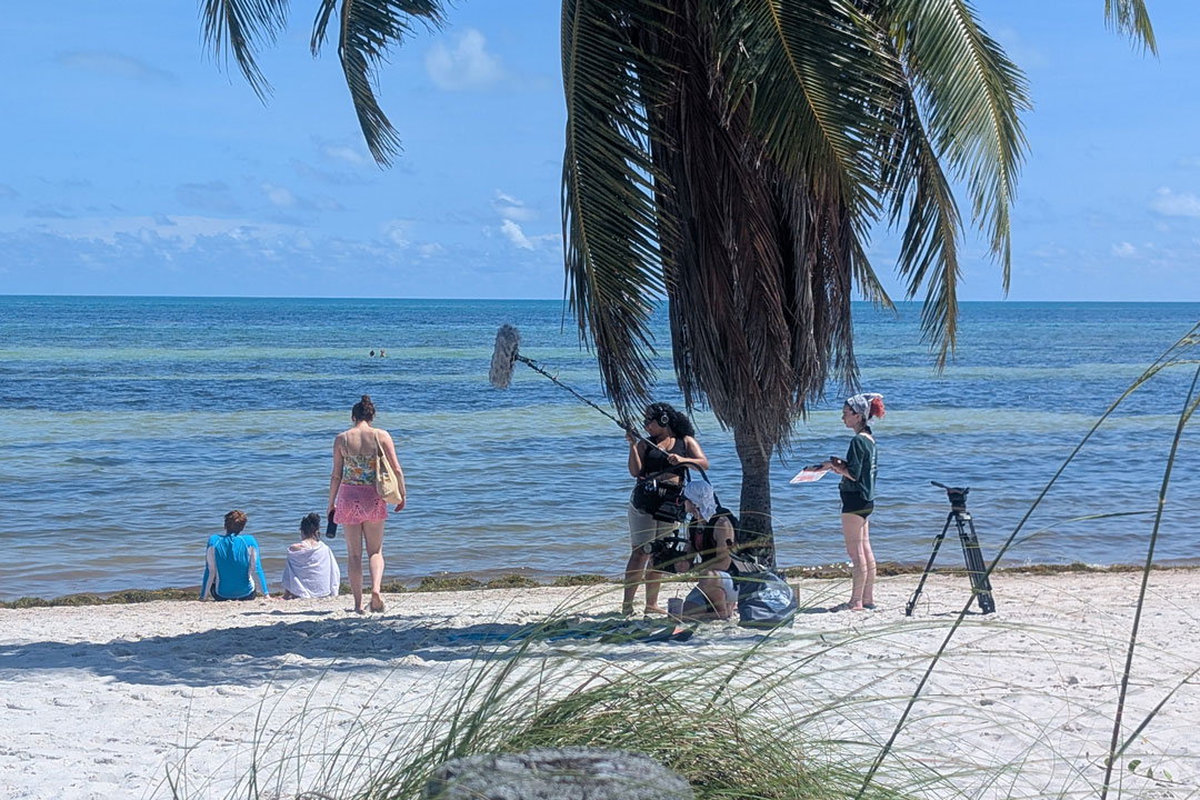 a large group of people stand ona. beach filming a movie scene