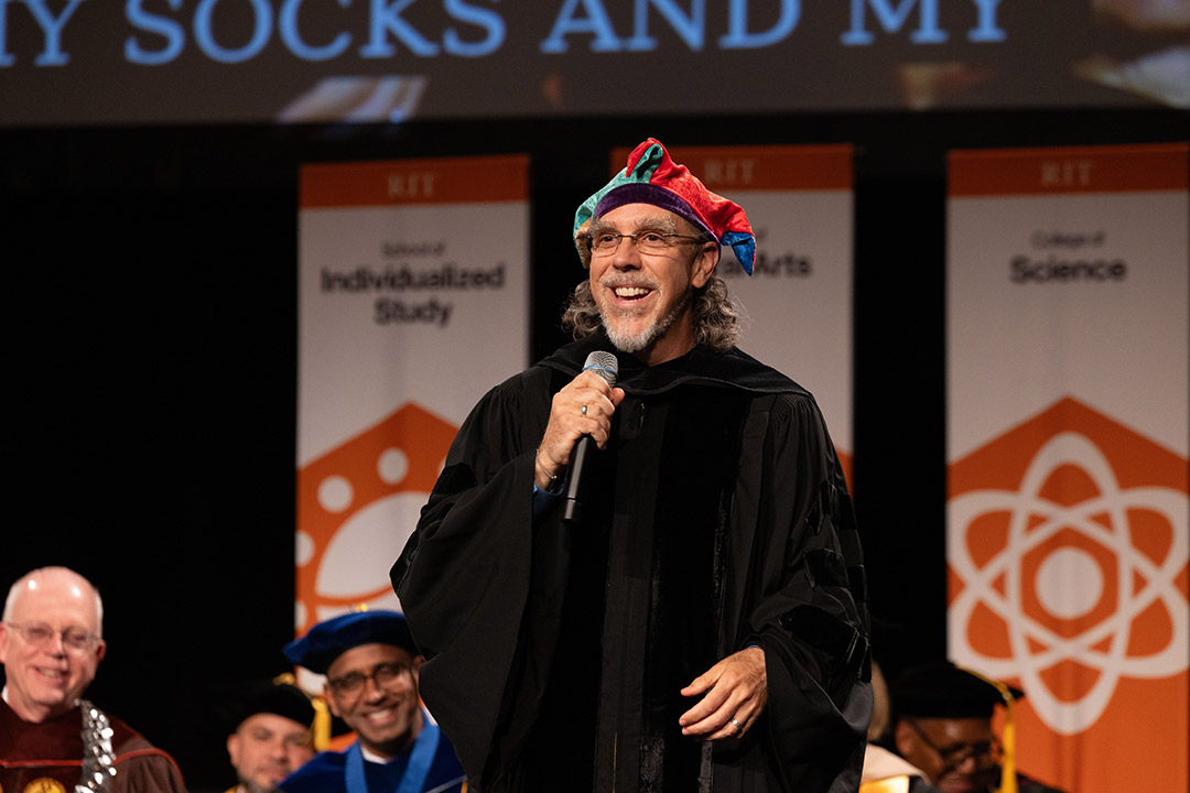 A person in academic regalia with a colorful jester hat speaks into a microphone at a ceremony, with RIT banners in the background.