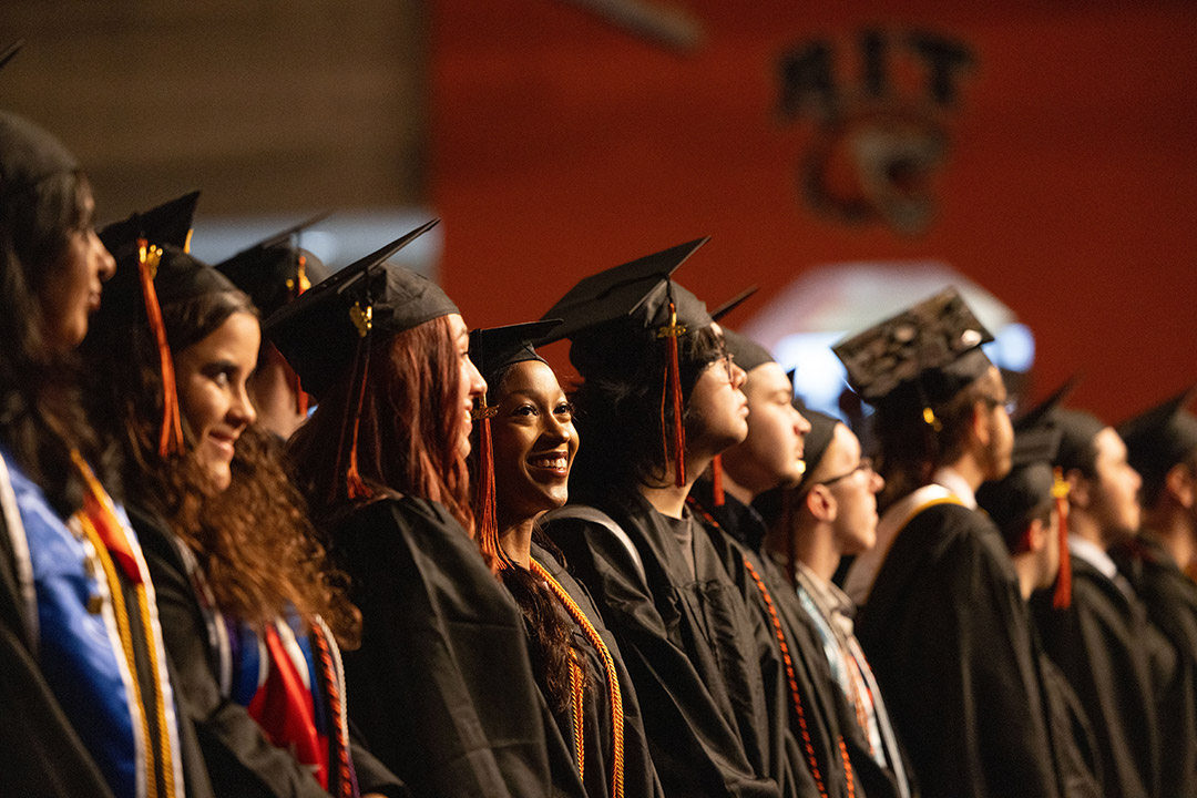 Graduates in caps and gowns at a commencement ceremony, with one smiling woman highlighted.