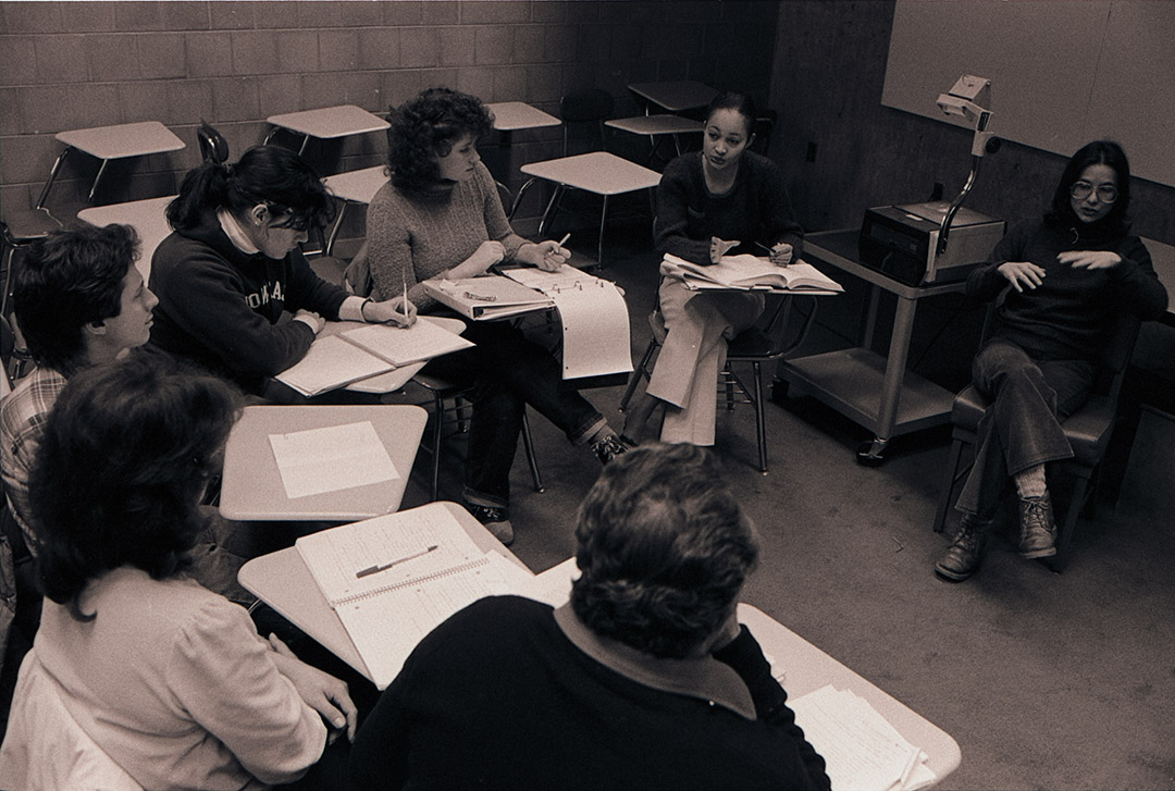 a brown and white photo of students sit in a classroom circle, taking notes and discussing with an instructor.